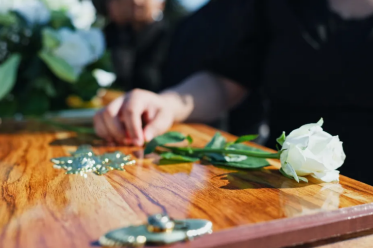 biała róża położona na drewnianej trumnie podczas ceremonii pogrzebowej, symbolizująca ostatnie pożegnanie, pamięć o zmarłej osobie, żałobę i godną oprawę uroczystości pogrzebowej